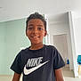 black_tshirt, boy, bright_lighting, casual_clothing, chair, child, curly_hair, doorway, floor, green_wall, happy, indoor, modern_room, nike_logo, person, portrait, smiling, standing, white_wall, young