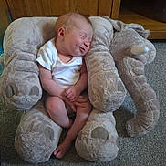 Skyla is registered to the contest to win money with this photo: baby, infant, smiling, plush, elephant, stuffed_animal, soft_toy, carpet, wooden_furniture, cozy, indoor, child, white_onesie, cute, sleepy, resting, small, adorable, peaceful, hands