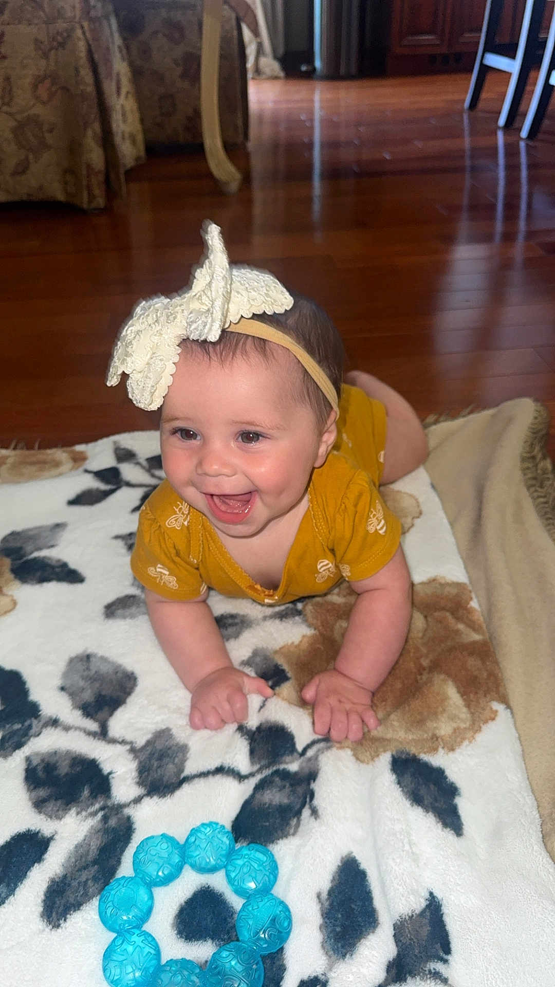 Ava is registered to the contest to win money with this photo: baby, child, smiling, headband, yellow_clothing, blanket, toy, blue_toy, floor, wooden_floor, indoor, happy, infant, cute, person, face, hands, crawling, home_interior, furniture