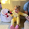 backpack, casual, child, floor, happy, home, indoor, light, minnie_mouse, person, pink_crocs, playful, plush_toy, shadow, sitting, smile, toddler, toy, wall, yellow_clothing
