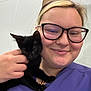 Tortellini is registered to the contest to win money with this photo: animal, black_cat, cat, closeup, cute, face, friendly, glasses, hand, holding, indoor, kitten, nose_piercing, person, pet, purple_clothing, short_hair, smile, veterinarian, young_adult