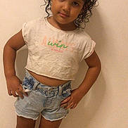 Aya a rejoint le concours — aidez-le/la à gagner de superbes lots ! child, curly_hair, standing, tshirt, denim_shorts, barefoot, indoor, tile_floor, confident, pose, casual_clothing, young, person, smiling, cute, portrait, wall, hands_on_hips, kid, fashion