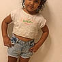 child, curly_hair, standing, tshirt, denim_shorts, barefoot, indoor, tile_floor, confident, pose, casual_clothing, young, person, smiling, cute, portrait, wall, hands_on_hips, kid, fashion