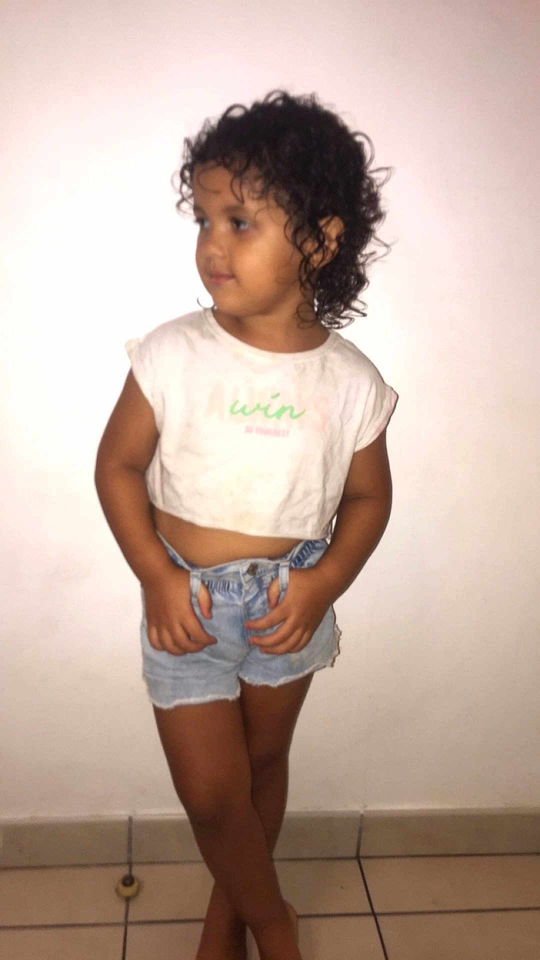 Aya a rejoint le concours — aidez-le/la à gagner de superbes lots ! child, curly_hair, denim_shorts, white_tshirt, standing, indoor, tiled_floor, wall, casual_clothing, portrait, barefoot, young, looking_away, hands, smile, shorts, cute, skin, floor_tile, posing