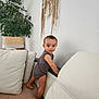 baby, child, indoor, curious, smiling, standing, carpet, couch, white_leather, plant, lamp, dreamcatcher, wall, home_decor, furniture, person, clothing, portrait, cute, happy