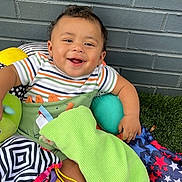 Princeton is registered to the contest to win money with this photo: baby, smiling, child, blanket, toys, cushion, colorful, striped_clothing, grass, brick_wall, happy, infant, playtime, indoors, comfort, person, cute, small_child, sitting, fabric