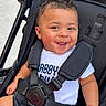 Princeton is registered to the contest to win money with this photo: baby, smiling, stroller, child, happy, face, clothing, toy, sitting, colorful, cute, infant, outdoor, person, black, white_shirt, blue_shorts, secure, playful, bright