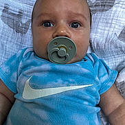 Taio is registered to the contest to win money with this photo: baby, pacifier, blue_shirt, tie_dye, infant, face, eyes, blanket, clothing, lying_down, cute, portrait, child, young, skin, head, expression, indoors, person, closeup