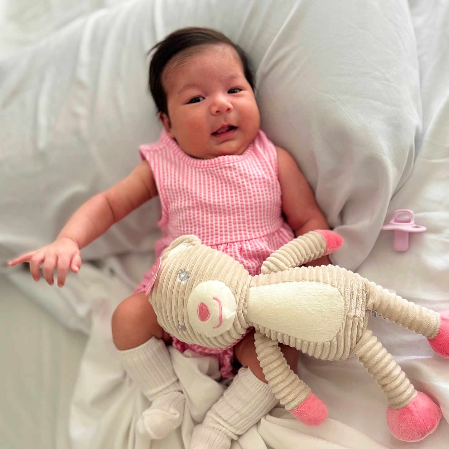 Lariany is registered to the contest to win money with this photo: baby, baby_girl, blanket, child, comfort, cozy, cute, happy, indoors, infant, pacifier, pink_dress, portrait, resting, sleepwear, smiling, socks, soft_toy, stuffed_animal, white_bed