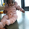 Jamilah is registered to the contest to win money with this photo: baby, child, infant, portrait, sitting, pink_outfit, knitted_clothing, sweater, hat, bow, mittens, booties, dark_skin, curly_hair, serious_expression, adult_hands, indoor, kitchen, countertop, window