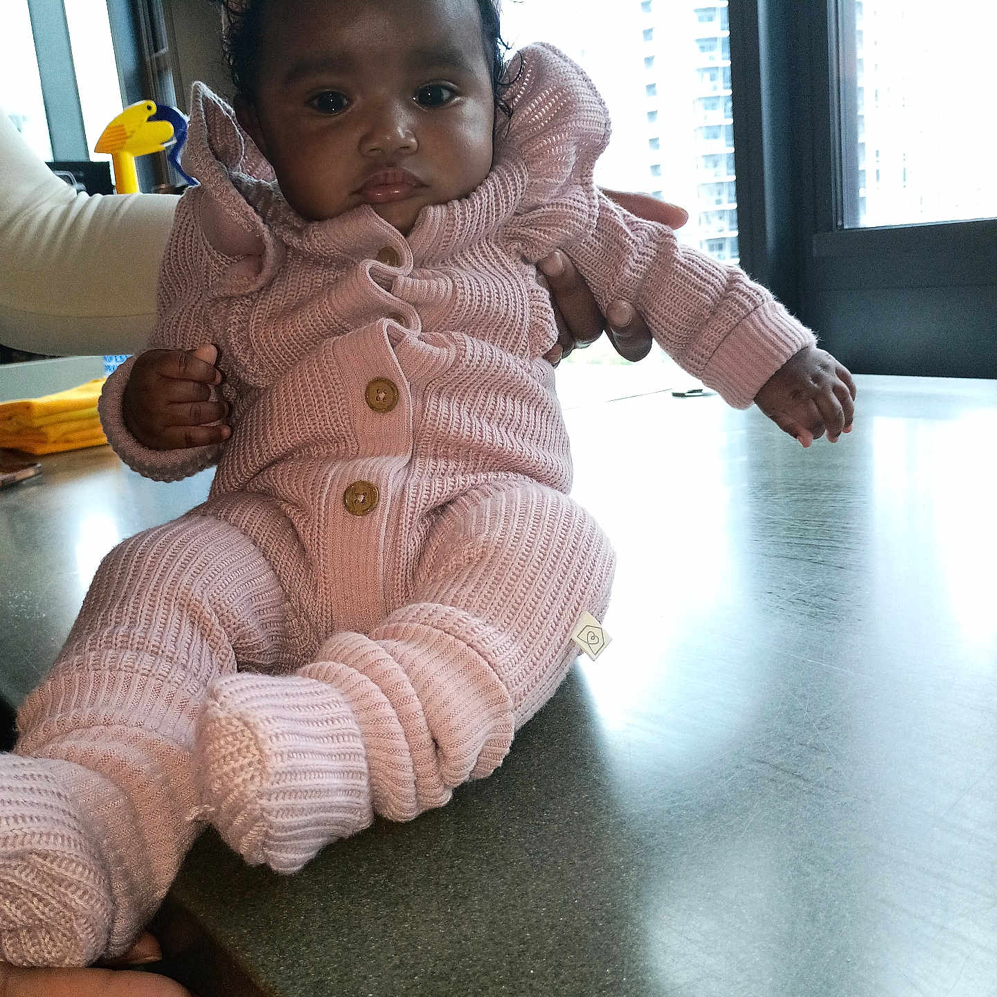 Jamilah is registered to the contest to win money with this photo: baby, child, infant, portrait, sitting, pink_outfit, knitted_clothing, sweater, hat, bow, mittens, booties, dark_skin, curly_hair, serious_expression, adult_hands, indoor, kitchen, countertop, window