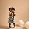 toddler, child, denim_shorts, denim_shirt, bucket_hat, clapping, balloons, studio, beige_background, sneakers, standing, happy, cute, portrait, indoor, fashion, smiling, person, footwear, playful