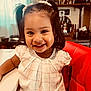 child, girl, smile, pigtails, hair_clips, red_chair, white_table, indoor, cute, happy, portrait, clothing, furniture, person, face, young_child, dressed, home, decor, cheerful