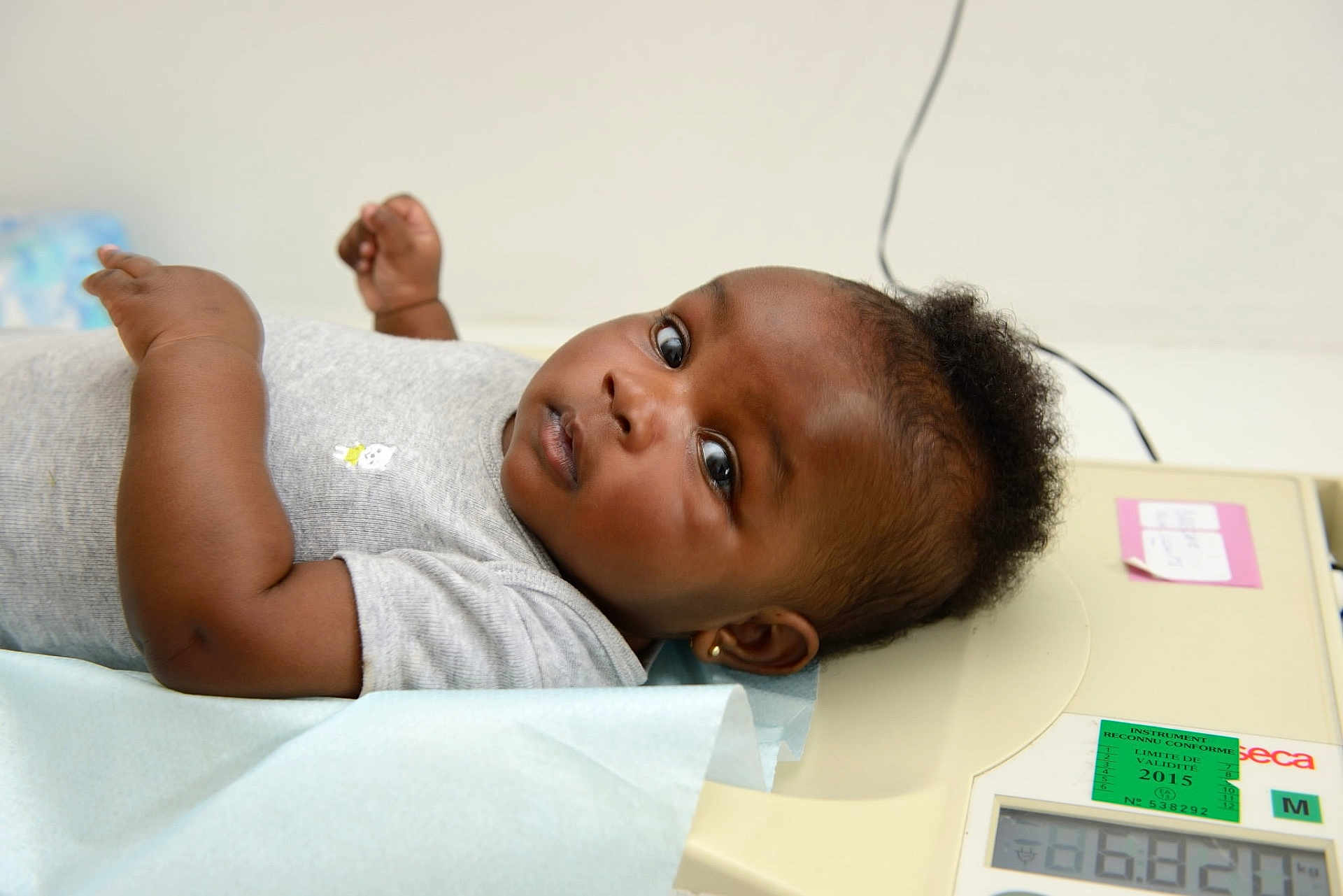 Elya-Victoria participe au concours pour gagner de l'argent avec cette photo : baby, infant, medical_scale, lying_down, gray_shirt, curly_hair, earrings, blue_paper, looking_at_camera, indoors, health_check, skin, child, person, portrait, cute, young, measurement, expression, resting