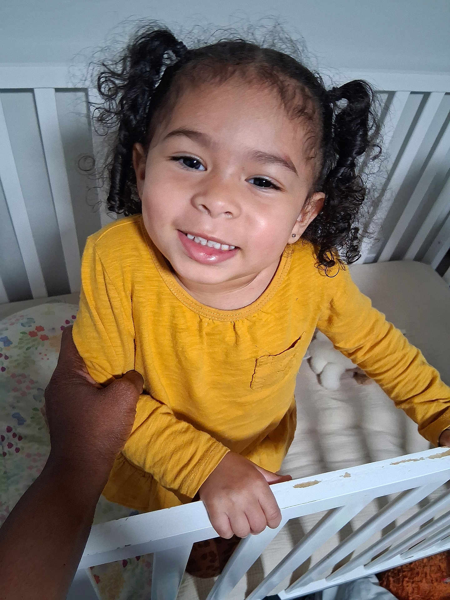 Leeah joined the competition — help win amazing prizes! toddler, child, smiling, yellow_shirt, crib, hand, person, indoor, curly_hair, bed, toy, furniture, happy, holding, portrait, baby, cute, skin, face, cozy