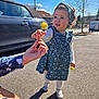 toddler, child, flower, dandelion, hand, outdoor, sunny, dress, headband, car, parking_lot, road, tree, curly_hair, person, footwear, shadow, blue_sky, sidewalk, interaction
