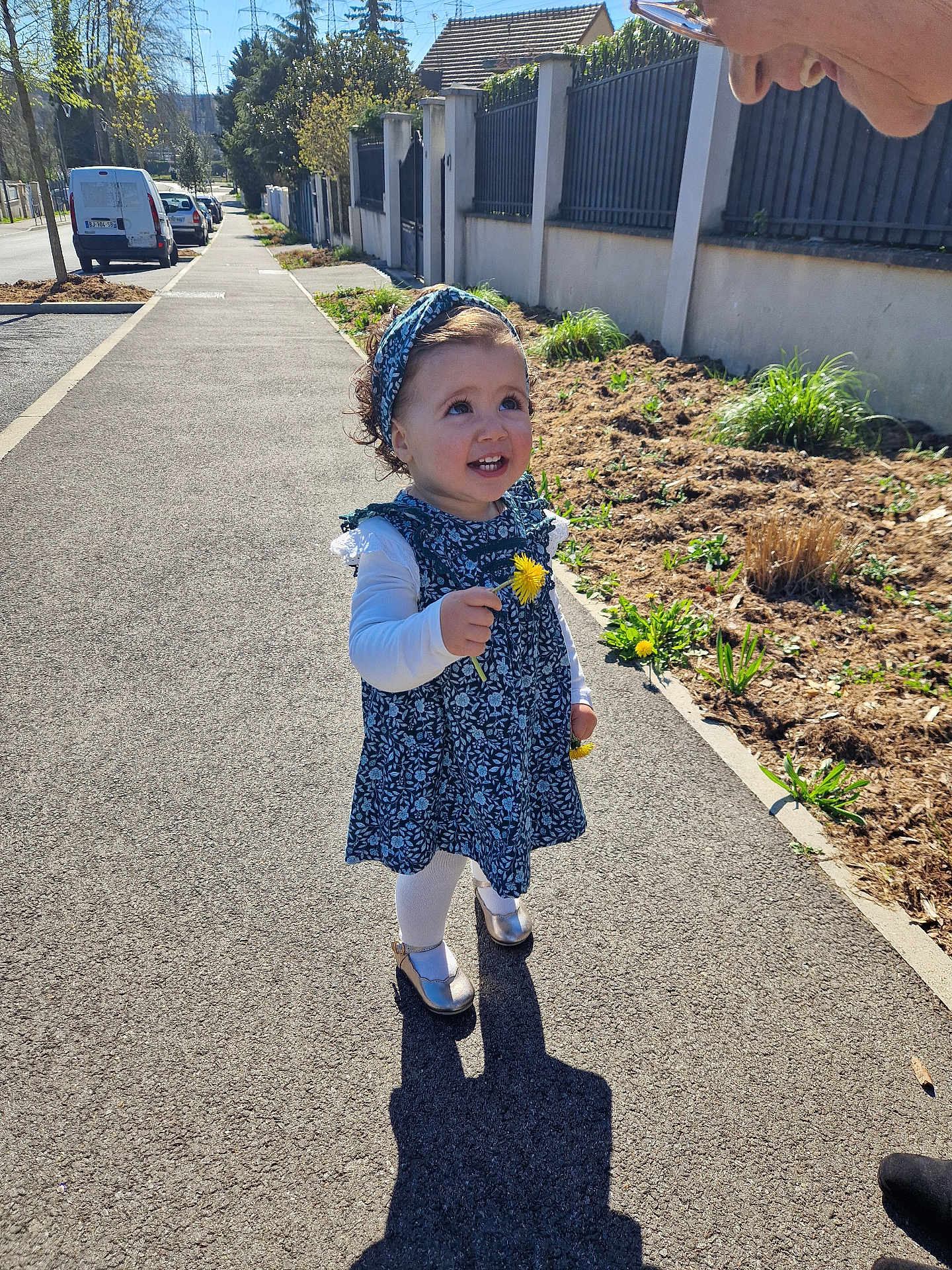 Chloé participe au concours pour gagner de l'argent avec cette photo : child, toddler, flower, dandelion, sidewalk, outdoor, sunlight, dress, headband, shoes, person, smiling, happy, curly_hair, nature, plants, road, fence, adult, shadow