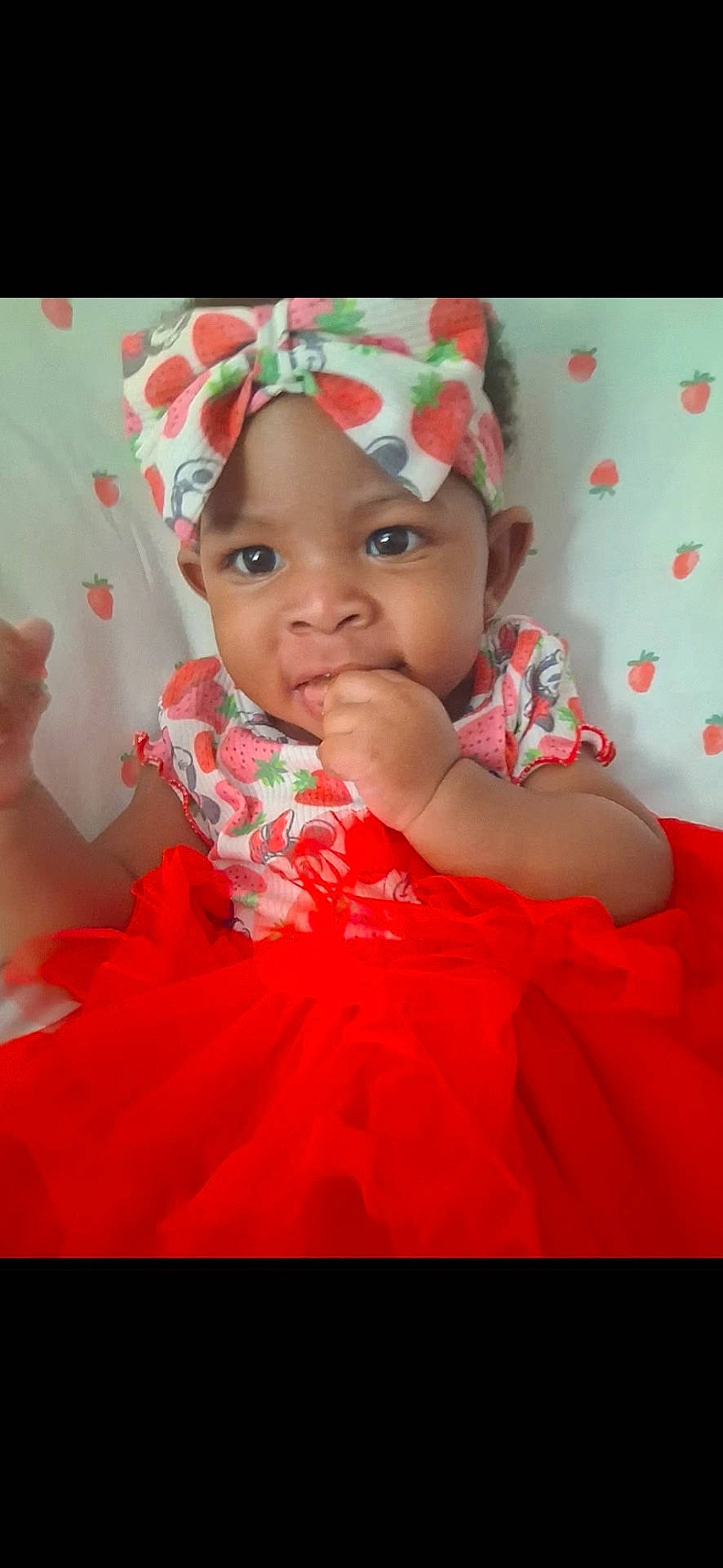 Saniyah is registered to the contest to win money with this photo: baby, background, child, clothing, colorful, cute, face, girl, hand_in_mouth, happy, headband, indoor, infant, person, portrait, red_skirt, sitting, smiling, strawberry_pattern, young_child