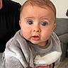 élio a rejoint le concours — aidez-le/la à gagner de superbes lots ! adult_holding, baby, blue_eyes, cheeks, child, close_up, couch, eyelashes, floor, gaze, grey_onesie, hand, indoor, infant, living_room, person, plush_clothing, portrait, soft_lighting, surprised_expression