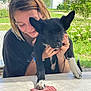 woman, dog, black_dog, white_paws, playing_cards, table, outdoor, greenery, smiling, happy, casual_clothing, daylight, nature, modern_building, cute, pet, friendship, relaxing, leisure, affection