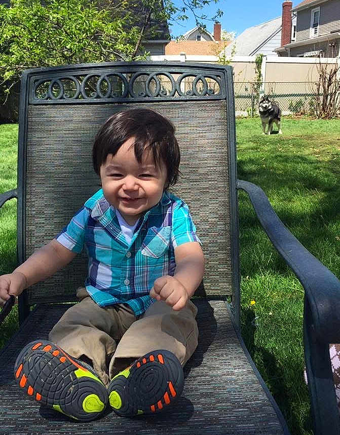 Christopher joined the competition — help win amazing prizes! toddler, child, smiling, outdoor, grass, chair, plaid_shirt, pants, shoes, dog, fence, backyard, sunny, happy, person, patio, greenery, casual_clothing, daytime, seated