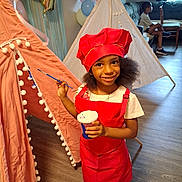 Bria is registered to the contest to win money with this photo: child, girl, chef_hat, red_outfit, paintbrush, cup, indoor, teepee, balloons, floor, couch, boy, window, curtains, wooden_floor, smile, playtime, party, decorations, shoes