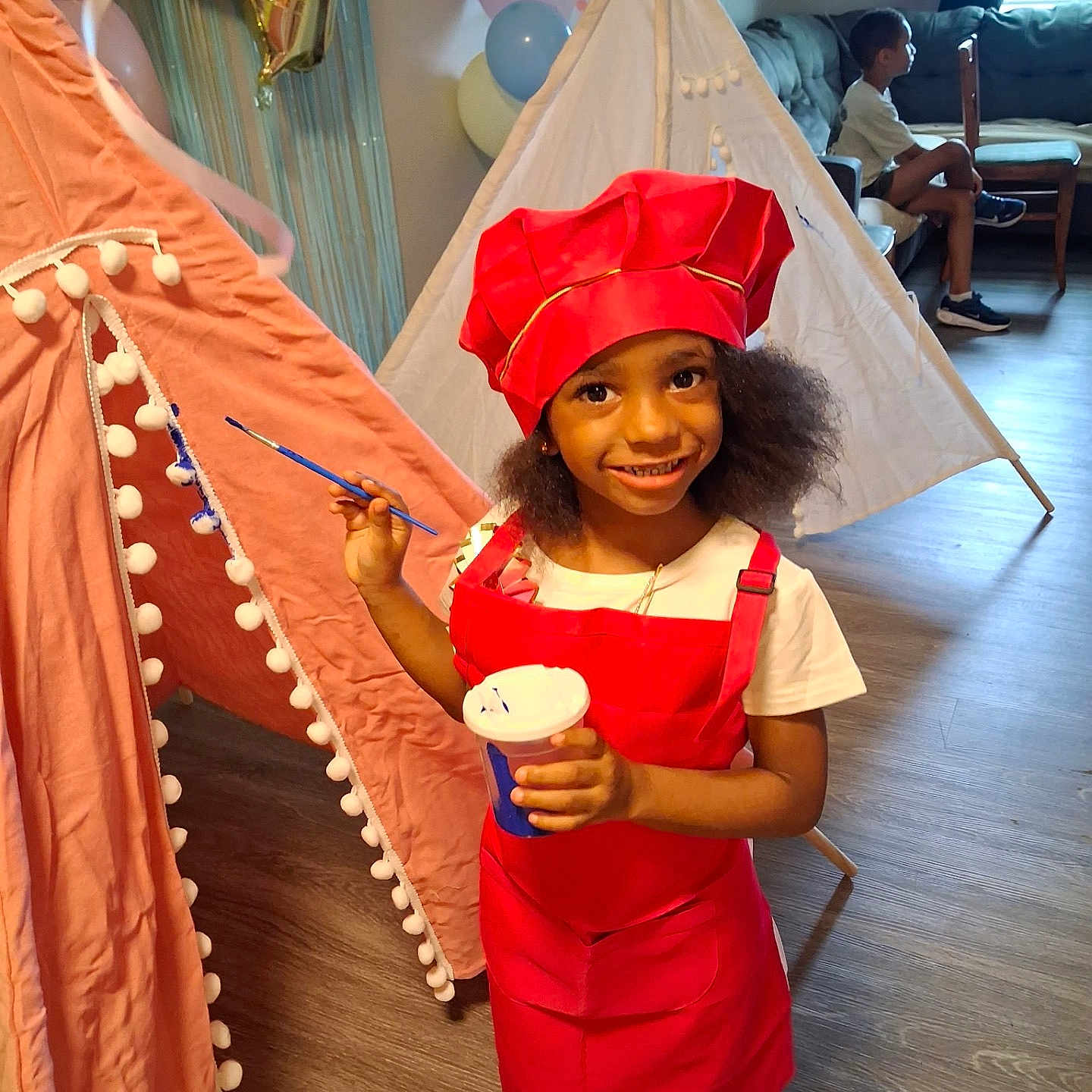 Bria is registered to the contest to win money with this photo: child, girl, chef_hat, red_outfit, paintbrush, cup, indoor, teepee, balloons, floor, couch, boy, window, curtains, wooden_floor, smile, playtime, party, decorations, shoes
