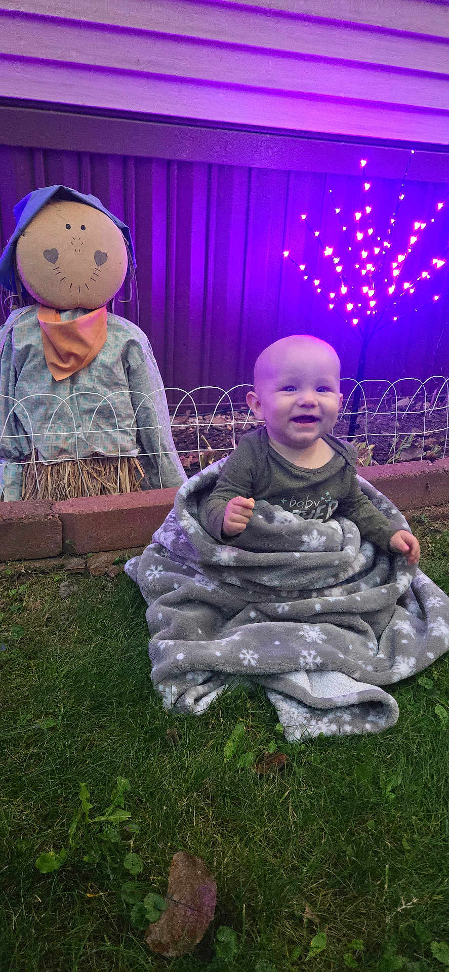 Hudson is registered to the contest to win money with this photo: baby, blanket, grass, scarecrow, purple_lights, smiling, outdoor, child, happy, face, person, fence, garden, night, clothing, head, toy, decoration, winter_theme, cozy