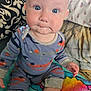 baby, child, blue_eyes, onesie, sitting, blanket, pillow, patterned_clothing, indoors, cute, infant, face, hand, footwear, colorful, cozy, expression, soft_fabric, head, person