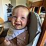 animal_prints, baby, bib, blue_eyes, brown_clothing, child, cozy, cute, food_stains, furniture, happy, high_chair, home, indoor, infant, natural_light, person, plant, portrait, smiling