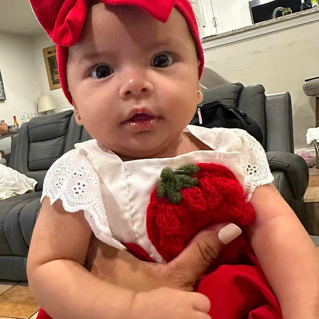Aislin is registered to the contest to win money with this photo: adorable, baby, child, closeup, couch, curious, cute, dress, furniture, hand, headband, holding, indoor, infant, living_room, person, portrait, red_bow, watch, white_wall