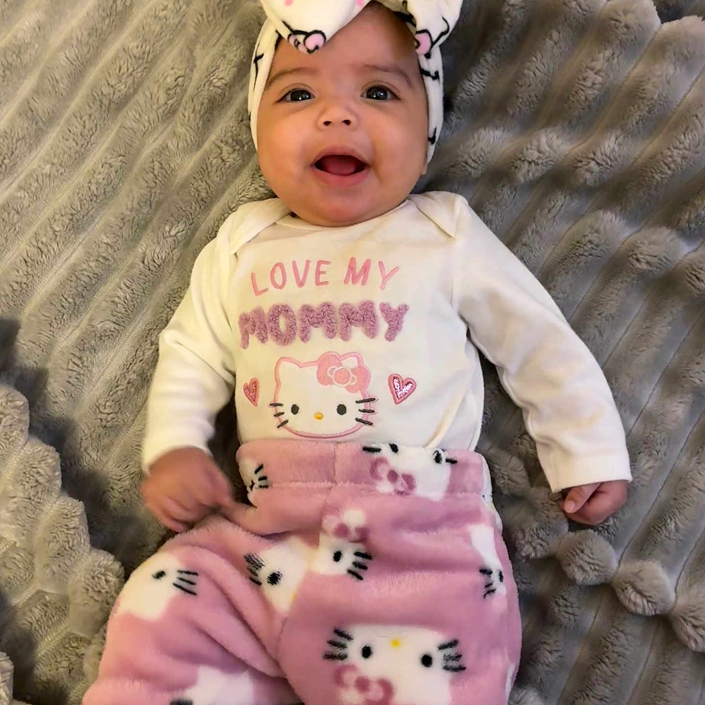 Kaylanna is registered to the contest to win money with this photo: baby, smiling, headband, hello_kitty, pink_pants, white_shirt, text_on_clothing, blanket, soft_texture, infant, happy, cute, child, lying_down, indoor, portrait, clothing, baby_girl, face, person
