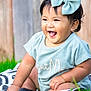 baby, child, smiling, laughing, bow, headwear, outdoor, grass, blanket, pattern, wooden_fence, happy, sitting, cute, toddler, apparel, short_sleeve, person, tongue_out, playful
