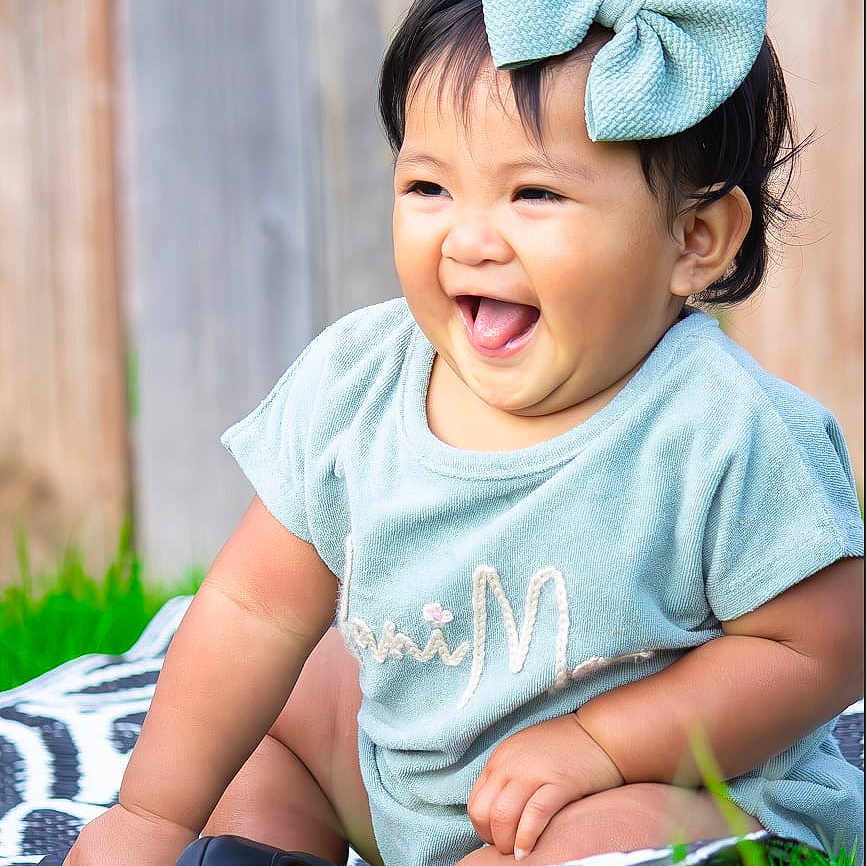 Alanimae is registered to the contest to win money with this photo: apparel, baby, blanket, bow, child, cute, grass, happy, headwear, laughing, outdoor, pattern, person, playful, short_sleeve, sitting, smiling, toddler, tongue_out, wooden_fence