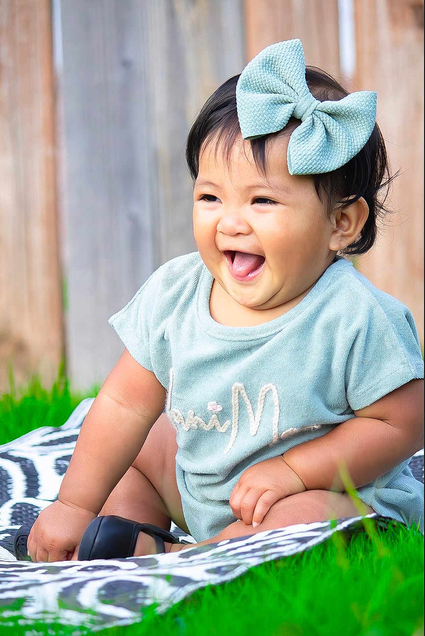 Alanimae is registered to the contest to win money with this photo: baby, child, smiling, laughing, bow, headwear, outdoor, grass, blanket, pattern, wooden_fence, happy, sitting, cute, toddler, apparel, short_sleeve, person, tongue_out, playful