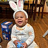 Ailani is registered to the contest to win money with this photo: accessory, baby, bunny_ears, chairs, child, cute, floor, happy, home, indoor, infant, lemon_pattern, onesie, person, playful, smiling, table, tablecloth, toy, wooden_floor