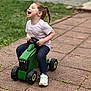 Macie is registered to the contest to win money with this photo: child, laughing, toy_tractor, braided_hair, outdoor, pavement, grass, casual_clothing, white_shoes, play, joy, happy, person, green_toy, side_view, daylight, young_child, fun, park, smile