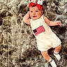 Brandi is registered to the contest to win money with this photo: baby, infant, smiling, romper, headband, bow, american_flag, socks, lying_down, gray_rug, soft_texture, cute, happy, child, indoors, portrait, one_person, skin, face, hand
