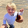 child, blonde_hair, white_shirt, jeans, blue_shoes, pumpkin_basket, hand_holding, outdoor, concrete, sunlight, smiling, person, daylight, casual_clothing, young_child, holding_basket, playful, toddler, happy, bright