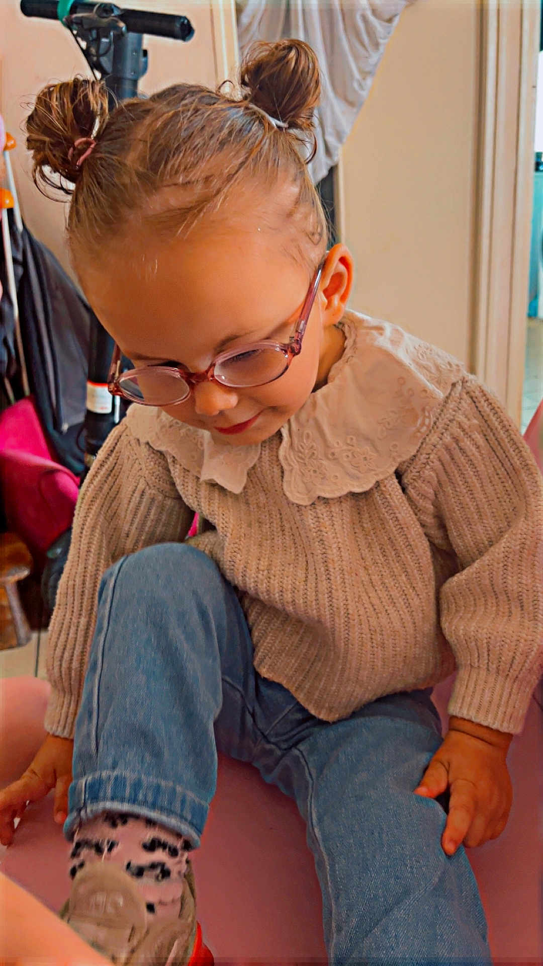 Larya participe au concours pour gagner de l'argent avec cette photo : child, glasses, hair_buns, sweater, lace_collar, jeans, socks, indoor, sitting, cute, smiling, young_child, portrait, cozy, casual_clothing, hand, footwear, home, furniture, playful
