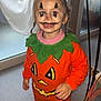 child, face_paint, pumpkin_costume, halloween, indoors, window, curtain, braids, sneakers, floor, smile, orange_clothing, green_collar, person, festive, holiday, costume, cute, standing, young