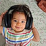baby, bed, black_headphones, child, colorful_clothing, comfort, cozy, cute, face, floral_sheet, hand, happy, headphones, indoor, infant, lying_down, person, smiling, striped_shirt, young_child
