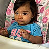 baby, blue_shirt, child, clothing, curious, cushion, dark_hair, expression, face, floral_pattern, hands, high_chair, indoor, infant, person, pink_flowers, portrait, sitting, toddler, young_child
