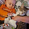 baby, cat, infant, person, adult, hand, orange_shirt, grandpa_shirt, pet, animal, cuddling, playful, motion_blur, indoor, blanket, couch, skull_pattern, text_on_clothing, family, portrait