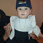 Briken is registered to the contest to win money with this photo: baby, child, infant, cap, hat, gold_chain, white_shirt, black_pants, sitting, indoor, person, hand, holding, face, wide_eyes, cute, fashion, portrait, expression, adult_hand