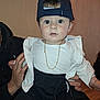 baby, child, infant, cap, hat, gold_chain, white_shirt, black_pants, sitting, indoor, person, hand, holding, face, wide_eyes, cute, fashion, portrait, expression, adult_hand