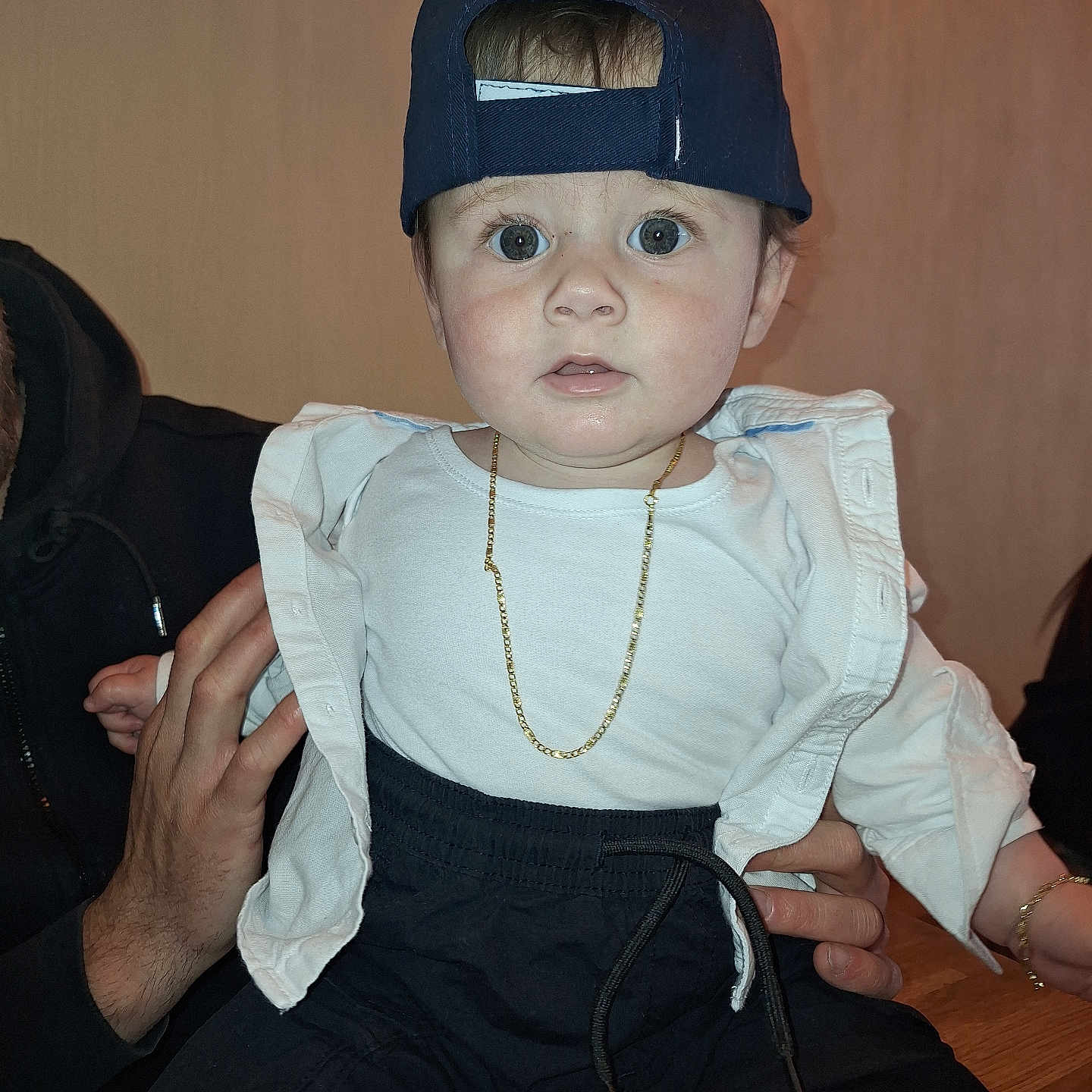 Briken is registered to the contest to win money with this photo: adult_hand, baby, black_pants, cap, child, cute, expression, face, fashion, gold_chain, hand, hat, holding, indoor, infant, person, portrait, sitting, white_shirt, wide_eyes