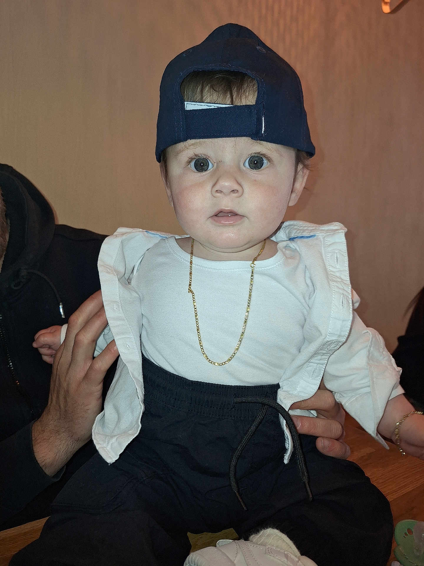 Briken is registered to the contest to win money with this photo: baby, child, infant, cap, hat, gold_chain, white_shirt, black_pants, sitting, indoor, person, hand, holding, face, wide_eyes, cute, fashion, portrait, expression, adult_hand