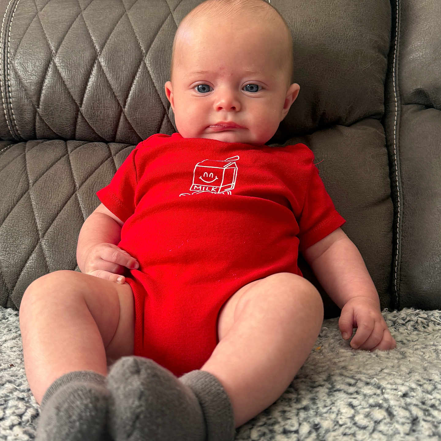 Tucker joined the competition — help win amazing prizes! baby, infant, red_onesie, milk_carton_graphic, gray_socks, quilted_couch, leather_furniture, blanket, blue_eyes, pout, sitting, indoors, cute, child, portrait, soft_lighting, texture, cozy, hand, footwear