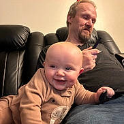 Osiris is registered to the contest to win money with this photo: baby, infant, child, adult, smiling, portrait, sitting, onesie, clothing, living_room, sofa, couch, furniture, phone, tablet, indoor, family, face, happy, home